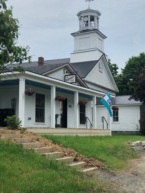 A one-story white building with a wrap-around porch with white columns that is used as a community gathering space in Albany, Vermont. A white clapboard church is in the background.
