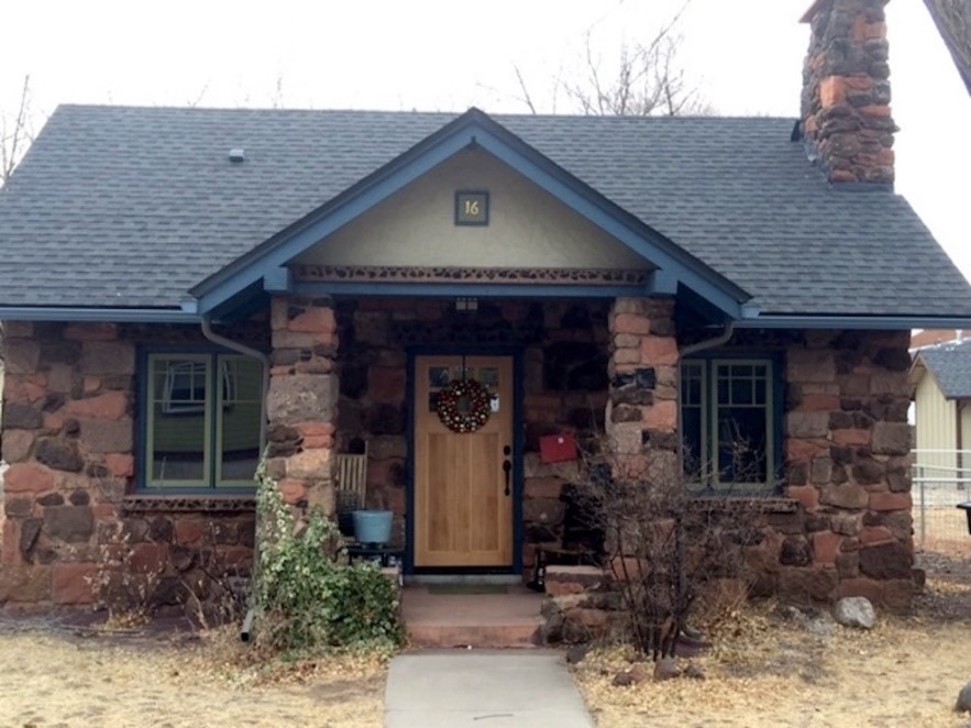 A small home built of local stone, with a stone chimney on the right and a gable-roofed entry porch. Located in Flagstaff, AZ.