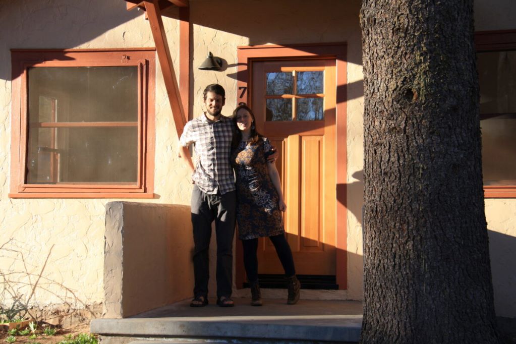 A man and woman standing in front of their front door. Bright sunlight highlights their smiles as new homeowners.