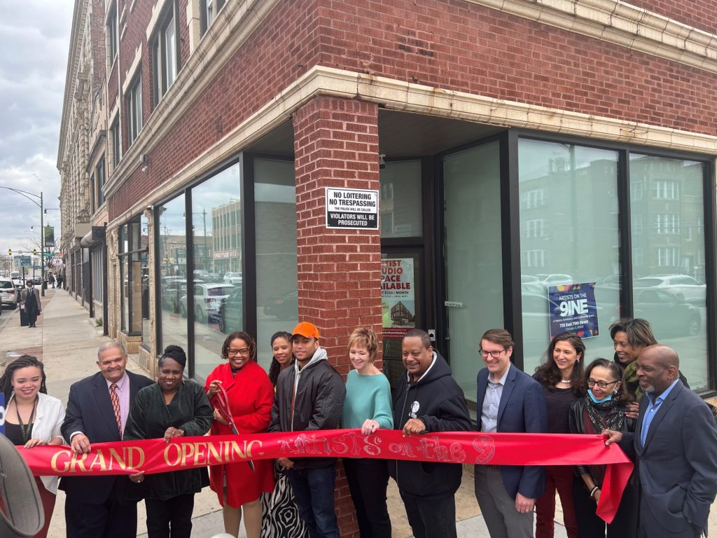 A group of people standing in front of a red brick building, getting ready to cut a large red ribbon at a ribbon cutting event.