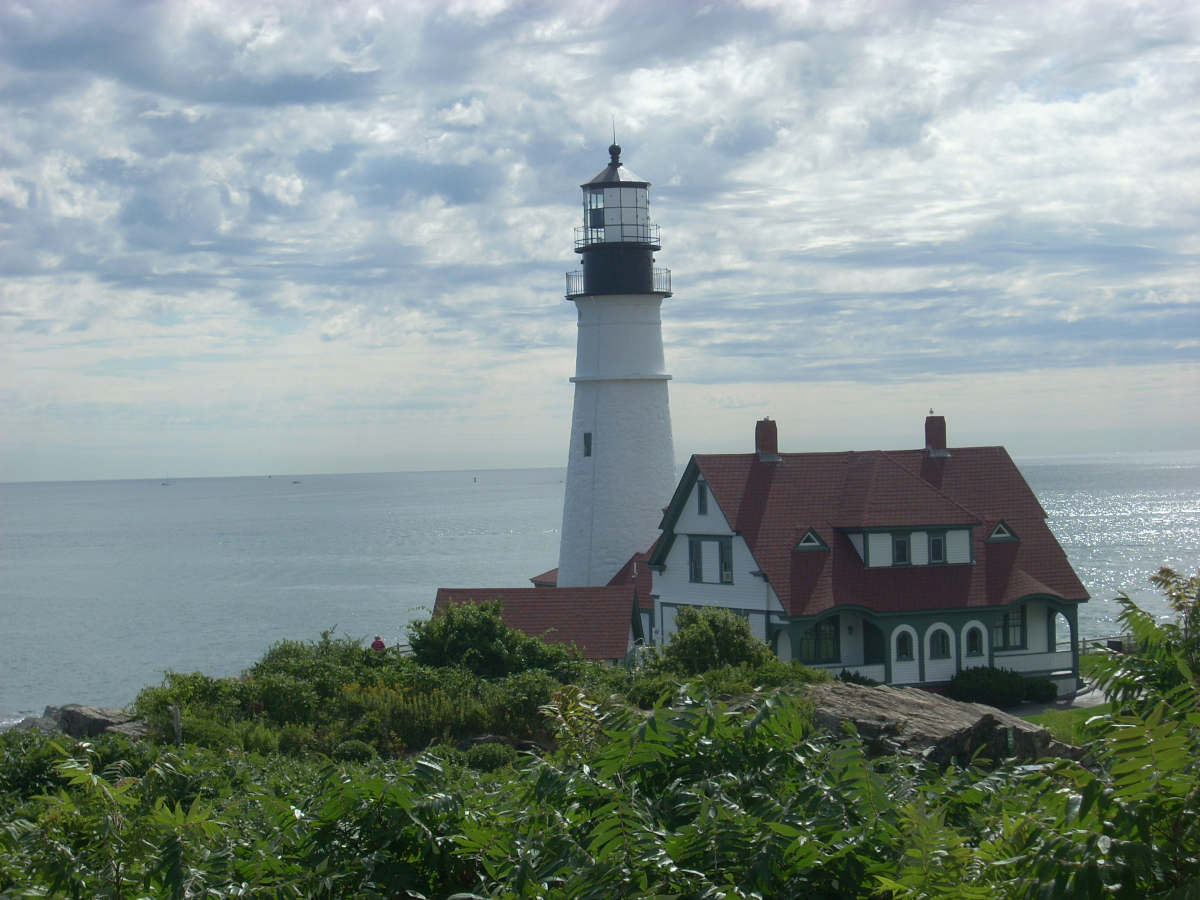 A decorative photograph of a lighthouse in Maine with the ocean behind it. The lighthouse is white with a black band near the beacon. The keeper's house's is white with green trim and a red roof. 