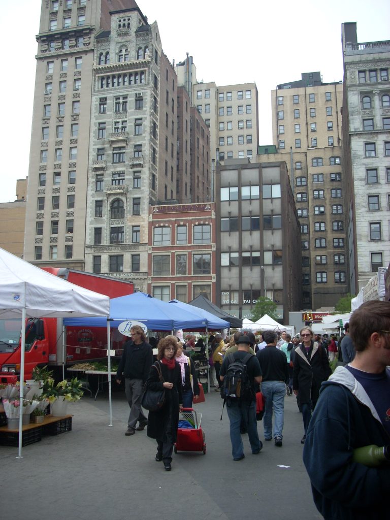 A busy scene in New York City with people shopping at a farmers market with tall buildings in the background.