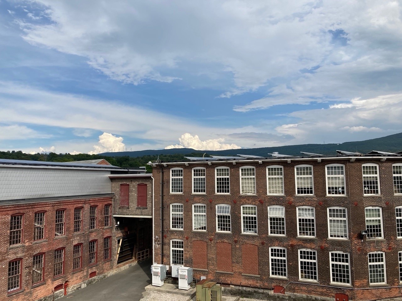 Photo of historic brick mill complex reused as an art museum. Solar panels are on the roof and along the mountain ridge in the distance, wind turbines are visible.